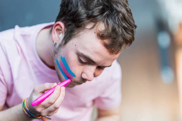 IMAGO Creative: Tag der Menschenrechte am 10. Dezember Man drawing colorful pink and blue stripes of transgender flag on cheek while standing in light room on blurred background HodeiUnzueta_TransgenderCallingForRights_45.jpg Copyright: xHodeixUnzuetax Model Released