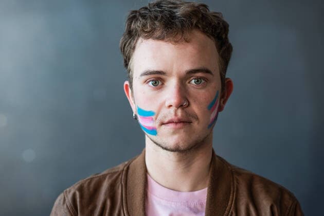 IMAGO Creative: Tag der Menschenrechte am 10. Dezember Joyful male with colorful painted blue and pink stripes representing transgender flag on cheek standing on gray background in light room HodeiUnzueta_TransgenderCallingForRights_52.jpg Copyright: xHodeixUnzuetax Model Released