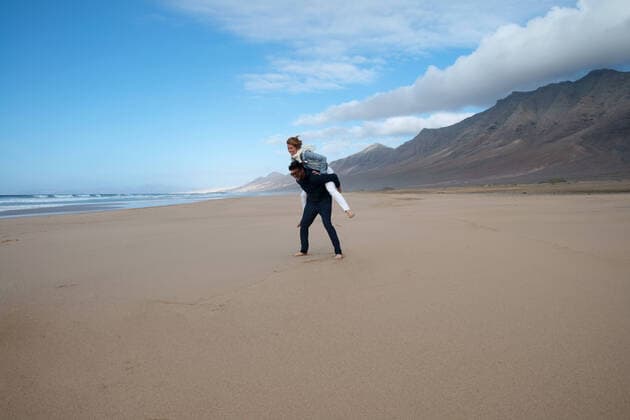 Creative Highlights Symbolbilder Full body of joyful barefoot woman having piggyback ride on boyfriend on wet sandy shore against hills on Fuerteventura island, Model released OscarDeDios_FuerteventuraMix_6.jpg Copyright: xOscarxDexDiosx MR