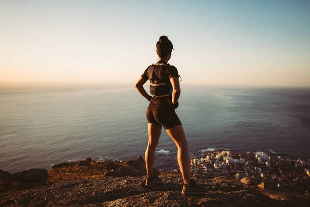 Creative Highlights Symbolbilder Back view of a woman in hiking sportswear standing on a cliff at sunset. Sportswoman looking at the ocean on the top of the mountain. Copyright: xArtemxVarnitsinxArtemxVarnitsinx