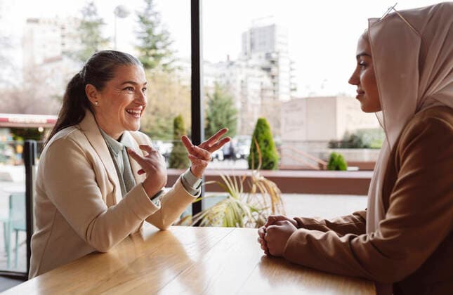 IMAGO Creative: Kollidierende Kulturen Smiling young businesswoman looking at colleague gesturing sitting at table in coffee shop model released, Symbolfoto, JCCMF05946