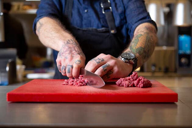 Cropped unrecognizable concentrated bearded male cook in uniform cutting fresh raw beef with knife on chopping board while cooking in light kitchen, Model released, Property released AlejandroMartinez_CastoKitchen_4.jpg Copyright: xAlejandroxMartinezx MR