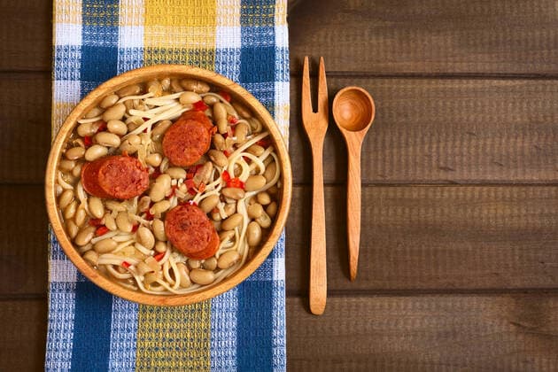 Overhead shot of traditional Chilean dish called Porotos con Riendas (English: beans with reins), made of cooked beans, linguine (flat spaghetti) and served with fried sausage, photographed on wood with natural light , 16386604.jpg, Chile, Chilean, cuisine, cooking, porotos, poroto, con, rienda, riendas, bean, reins, cooked, dish, meal, sausage, meat, chorizo, slice, spaghetti, pasta, flat, flattened, linguine, traditional, typical, food, nutrition, thick, stew, soup, pulse, vegetable, legume, wood, wooden, bowl, rustic, towel, fabric, cloth, kitchen, overhead, shot, copy, space, cutlery, fork, spoon, South, Latin, America, American, horizontal,