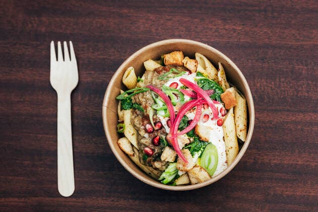 Top view of yummy pasta served in paper bowl with sauce and greens garnished with pickled onion and pomegranate seeds with croutons near plastic fork on wooden table HodeiUnzueta_BeachAfternoon_27.jpg Copyright: xHodeixUnzuetax Model Released