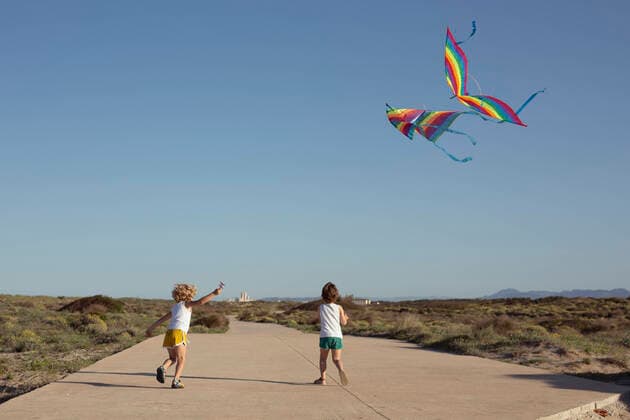 IMAGO Kreativ: Bruderschaft Full length of delighted little children in shorts walking on road and smiling while launching colorful kites against cloudless blue sky on sunny summer day SinuheGorris_EveningWithFriends_00016.jpg Copyright: xSinuhexGorrisx Model Released