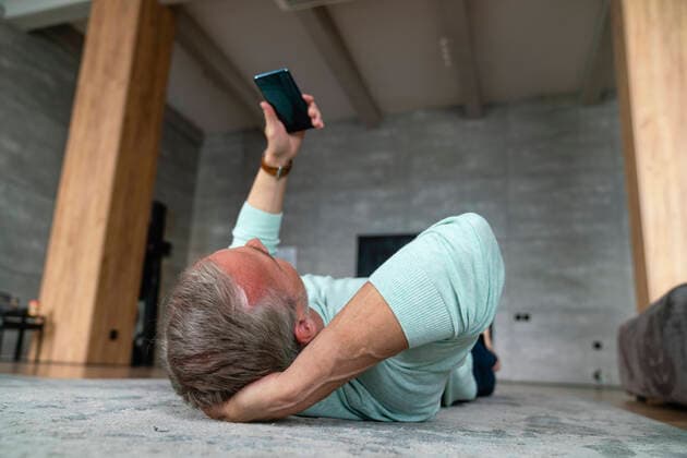 IMAGO Creative: Internationaler Tag der Architektur am 3. Oktober. Thema: Architektur zum Wohlfühlen Mature man with hand behind head using smart phone lying on carpet at home model released, Symbolfoto property released, VPIF06144
