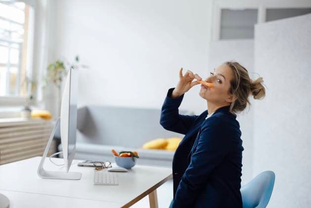 Playful businesswoman holding carrot on lips at desk in office model released, Symbolfoto property released, JOSEF10091