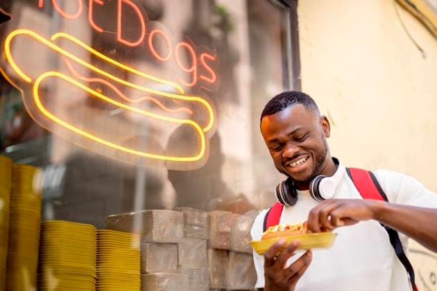 IMAGO Creative, Urban Lifestyle Happy young man eating hot dog leaning on restaurant window model released, Symbolfoto property released, OCMF02415