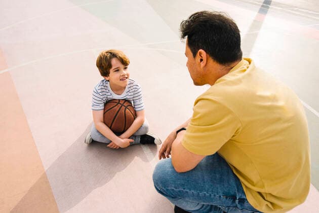 IMAGO Creative: 2022 Sport Favoriten IMAGO Creative: Lokalhelden - Jugendsportbildung Boy with basketball sitting in front of father at sports court on sunny day model released, Symbolfoto, MMPF00108