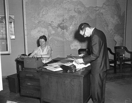 Trip to Europe: Arrival in Vienna, Austria 10:50AM; White House Army Signal Agency (WHASA) staff in Vienna. An unidentified woman sits behind a desk and an unidentified man stands in an unidentified building during White House Army Signal Agency (WHASA) preparations for a summit between President John F. Kennedy and Soviet Union Premier Nikita Khrushchev in Vienna, Austria. A large map of the world and a calendar are visible on the wall behind the woman. Copyright: xpiemagsx