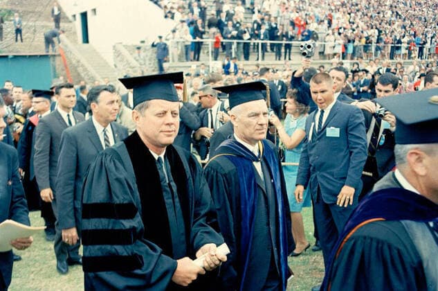 Trip to Western States: San Diego, California, arrival at Lindbergh Field, motorcade, and commencement address at San Diego State College. President John F. Kennedy (left) attends commencement exercises at San Diego State College. President of San Diego State College, Dr. Malcolm A. Love, stands right of President Kennedy. Standing at left (L-R): White House Secret Service agents, Gerald A. Jerry Behn (on far left edge of frame), Charlie Kunkel (in back), and Emory Roberts (behind President Kennedy). Aztec Bowl Stadium, San Diego State College, San Diego, California. Copyright: xpiemagsx