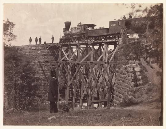 Bridge on Orange and Alexandria Rail Road, as Repaired by Army Engineers under Colonel Herman Haupt 1865 Andrew Joseph Russell American This view by Russell documents the engine Fire Fly testing the stability of a new wooden trestle bridge built quickly by the United States Military Railroad engineers to replace a masonry bridge destroyed by the Confederates. Atop the box car trailing the engine is a pair of armed sentries; others are on the rail tracks and bridge foundation. The top-hatted figure in the foreground is unidentified, but he may be the photographer himself or a civilian railroad engineer working for General Herman Haupt, commander of the United States Military Railroad. President Abraham Lincoln was so impressed with Haupt™s Copyright: xpiemagsx Metbot-31959