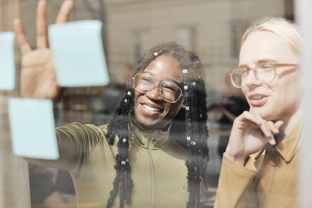 Creative Highlights Symbolbilder Smiling female entrepreneur explaining strategy to non-binary colleague on adhesive note over glass window in office Sweden, Stockholm, model released, property released Copyright: xMaskotx , MA94247 MA94247