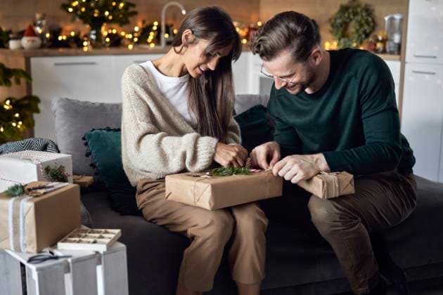 Smiling woman with man wrapping Christmas gift sitting on sofa at home model released, Symbolfoto property released, ABIF01717