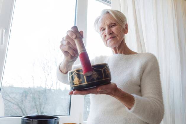 Smiling senior woman with singing bowl standing by window at home model released, Symbolfoto property released, OSF00216