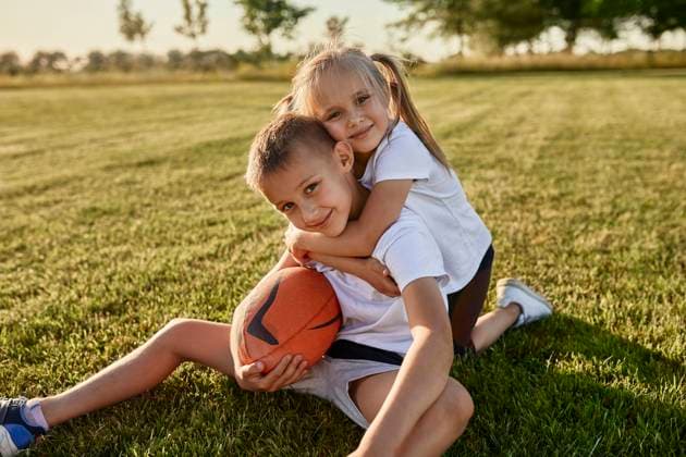 Smiling blond girl hugging brother sitting with rugby ball at sports field model released, Symbolfoto, ZEDF04688