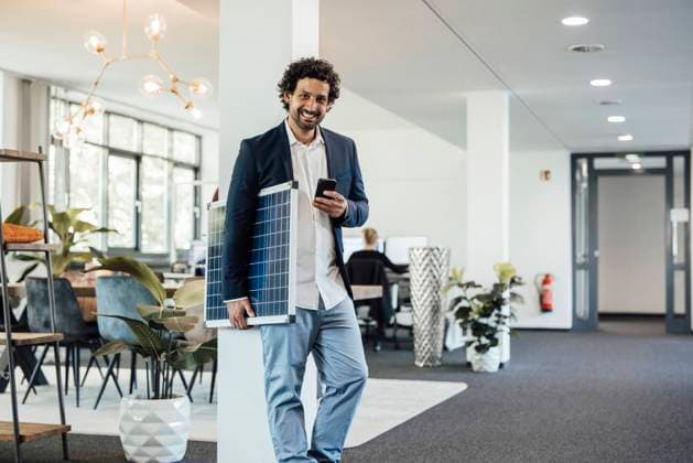 IMAGO Creative: Klimawandel - Klimabewusst Smiling businessman with smart phone and solar panel in office model released, Symbolfoto property released, JOSEF11728