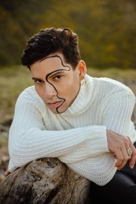 Young trendy guy with curvy ornament on face leaning on stone and looking at camera on sunny day in nature, Model released ErikaAnes_ManOnTheBeach_37 Copyright: xErikaxAnesx MR
