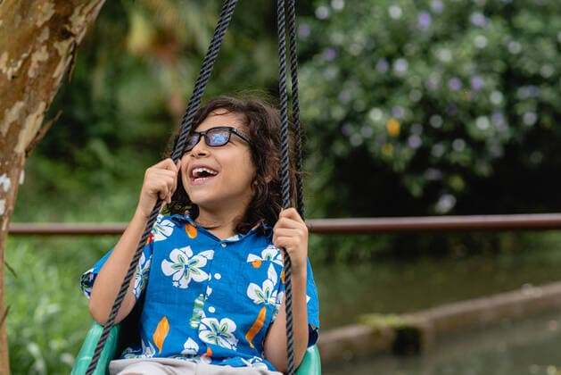 IMAGO Creative: Sommer, Sommertage im Garten Cheerful little boy with curly hair in casual clothes and eyeglasses sitting on swing and looking away in green park in daytime, Model released LeonardoBorges_FishingDay_13 Copyright: xLeonardoxBorgesx MR