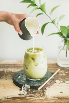 Iced matcha latte drink with milk pouring from pitcher Iced matcha latte drink in glass with coconut milk pouring from pitcher by woman s hand, white wall and plant branches at background. Summer refreshing beverage cold drink Copyright: xAnnaxIvanovax AI130718-4