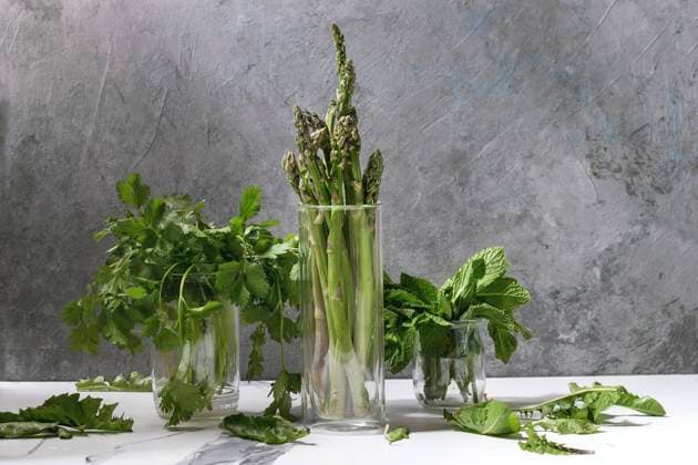 Asparagus ang greens Raw uncooked organic green asparagus, bunch of coriander herbs and fresh mint in glass jars on white kitchen table with grey texture wall as background. Copy space. Copyright: xNatashaxBreenx NB170518-49