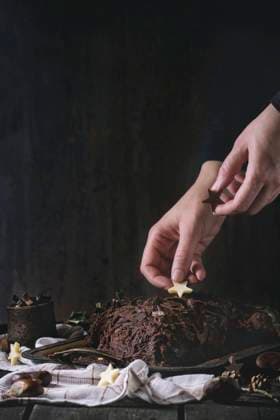 Decorating of christmas chocolate yule log Decorating process of homemade Christmas chocolate yule log by woman s hands with chocolate stars over old wooden table with holly branch and chestnuts. Dark rustic style. With retro filter effect Copyright: xNatashaxBreenx NB271016-3