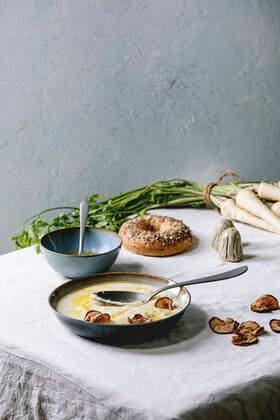 Parsnip soup with butter sauce Parsnip cream soup in ceramic bowl with butter sauce, sun dried pears, bundle of fresh parsnip, bagel bread and herbs on kitchen table with white tablecloth Copyright: xNatashaxBreenx NB300919-8