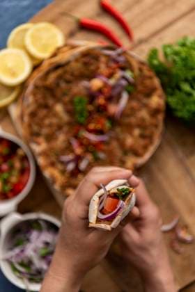 Hands holding a rolled up lahmacun. more lahmacun portions and condiments like red onion salad, tomato salad, lemon slices, parsley and chili pepper accompany. Copyright: xYusufxRizaxGunaydinx YG100720-13