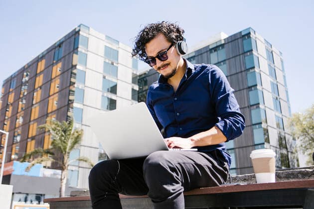 From below positive male entrepreneur text messaging on laptop sitting on bench and takeaway coffee on street, Model released MarcosCastillo_LatinBusiness_50 Copyright: xMarcosxCastillox MR