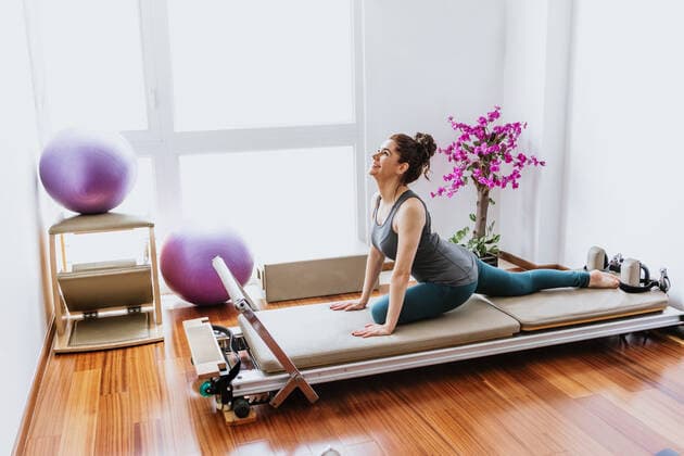 Side view of cheerful young female in activewear smiling while performing Ardha Kapotasana yoga pose on Pilates reformer in light room, Model released, Property released MarcosCastillo_Wellness_25 Copyright: xMarcosxCastillox MR
