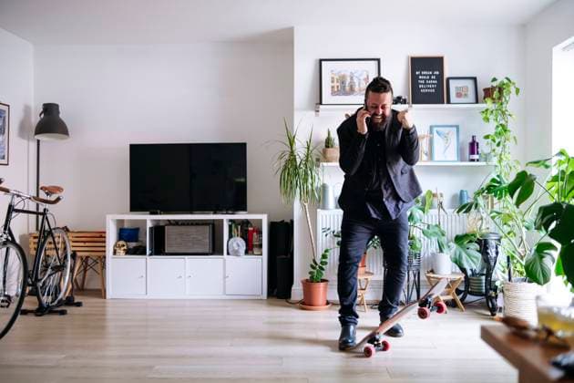 IMAGO Creative: Business, Innovative Technologie-Lösungen Cheerful businessman with skateboard talking on smart phone and clenching fist at home model released, Symbolfoto property released, ASGF02709