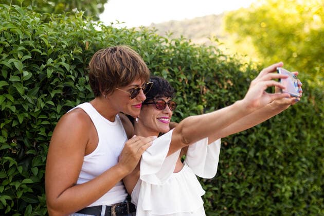 IMAGO Creative, Jahreszeit Sommer, Sommer in der Stadt Side view of stylish adult female friends standing near green shrubs in park and taking selfie on smartphone while spending summer weekend together PicuaEstudio_Lifestyle_18 Copyright: xPicuaxEstudiox Model Released