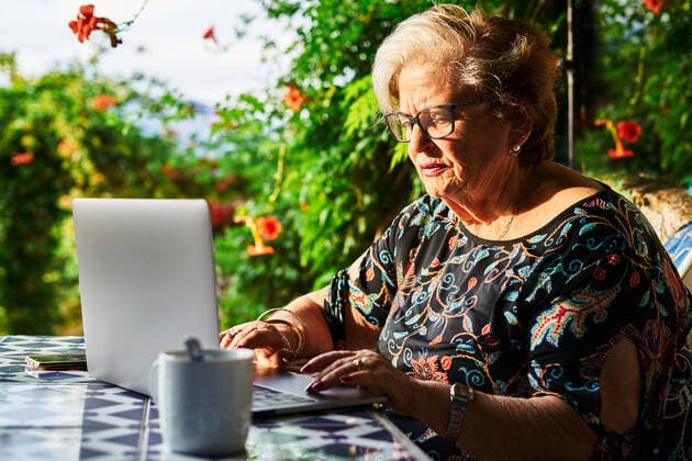 IMAGO Creative: Sommer, Sommertage im Garten Concentrated elderly lady in casual outfit and eyeglasses sitting in chair at table while surfing on netbook near cup on saucer and green plants with flowers in yard in daylight, Model released AdalbertoRodriguez_SeniorWomanUsingLaptop_12 Copyright: xAdalbertoxRodriguezx MR