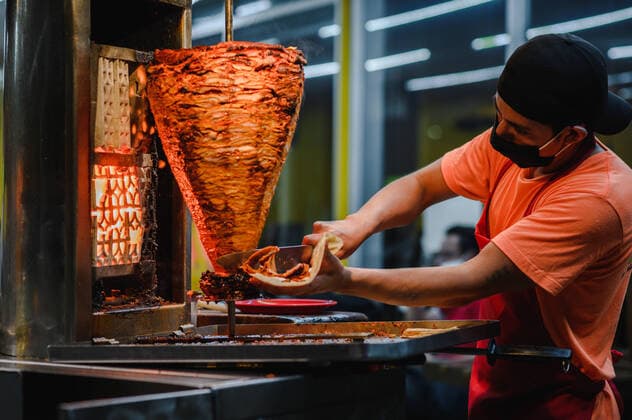 Side view ethnic male cook in cloth face mask with tortilla against vertical broiler with beef during COVID 19 pandemic, Model released, Property released HugoFuentes_MexicanTaco_8 Copyright: xHugoxFuentesx MR