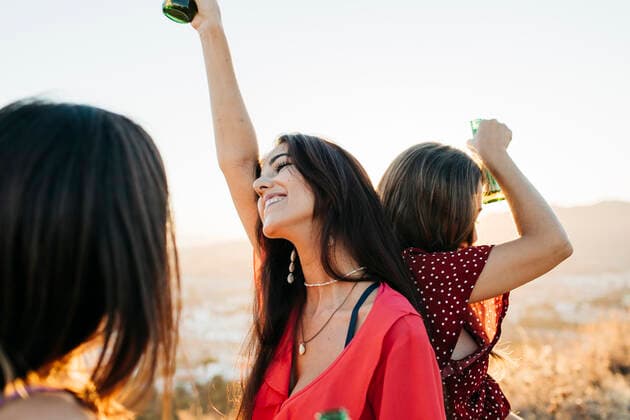 Delighted female friends in stylish outfits with bottles of alcohol beverages standing back to back while dancing on hilltop over town against cloudless sky in sunlight, Model released Lightsy_FriendsMountain_45 Copyright: xLightsyx MR