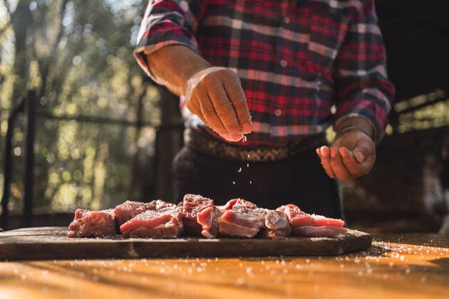 Crop unrecognizable man in checkered shirt putting salt on pieces of meat on wooden cutting board while preparing food in woods, Model released ReneStevens_ArgentinianBBQDance_14 Copyright: xRenexStevensx MR