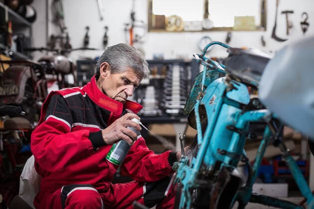 Side view of serious senior male master in workwear lubricating details of old retro motorbike with oil spray during repair work in garage OscarGimbal_SeniorMechanic_19 Copyright: xx Model Released
