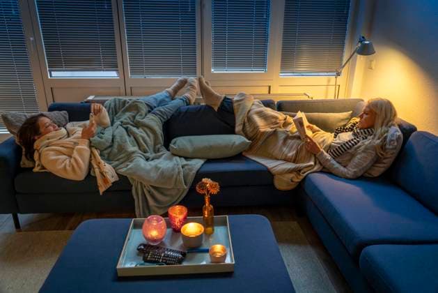 Symbolbild Energiesparen, kalter Winter, Energiekrise, kalte Wohnung, Mutter und Tochter liegen, warm angezogen auf dem Sofa, Kerzenlicht, Heizung auf ein Minimum an, wärmen sich die Füße am Heizkörper Energiesparen *** Symbol image energy saving, cold winter, energy crisis, cold apartment, mother and daughter lying, dressed warmly on the sofa, candlelight, heating to a minimum on, warm their feet on the radiator energy saving
