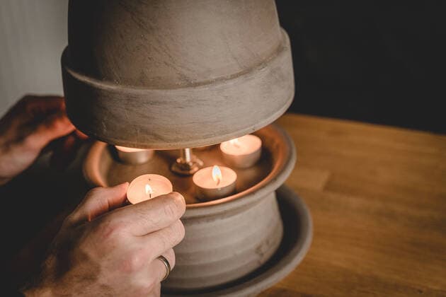 14 October 2022: young warmly dressed man warms his hands in a cold apartment without light on a tealight heater to save electricity and gas. homemade tealight stove, DIY heater made of plant pot and tealight candles *** junger warm angezogener Mann wärmt sich die Hände in einer kalten Wohnung ohne Licht an einer Teelichtheizung um Strom und Gas zu sparen. selbstgebauter Teelicht Ofen, DIY Heizung aus Pflanzentopf und Teelicht Kerzen