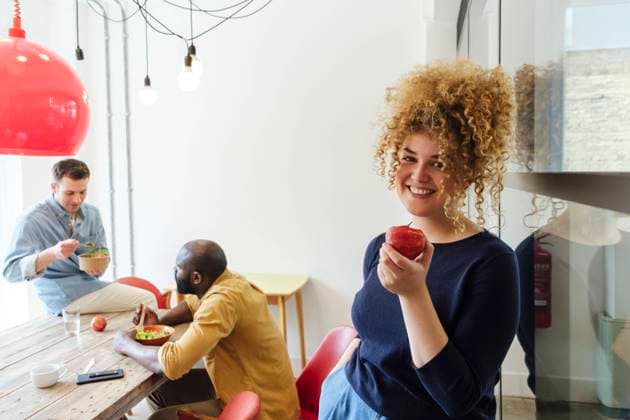 Smiling young woman eating apple in staff room leaning on wall model released, Symbolfoto property released, WESTF25053