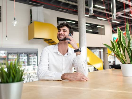 Smiling young bearded man in smart casual outfit standing near table and talking on smartphone while looking away in modern workspace AlbertMorcillo_ID3269_106754_027 Copyright: xAlbertxMorcillox Model Released