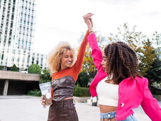Happy young multiracial girlfriends in street style outfits walking on city street near urban building and holding raised arms while looking at each other, Model released AlbertMorcillo_ID10549_319636_008 Copyright: xAlbertxMorcillox MR