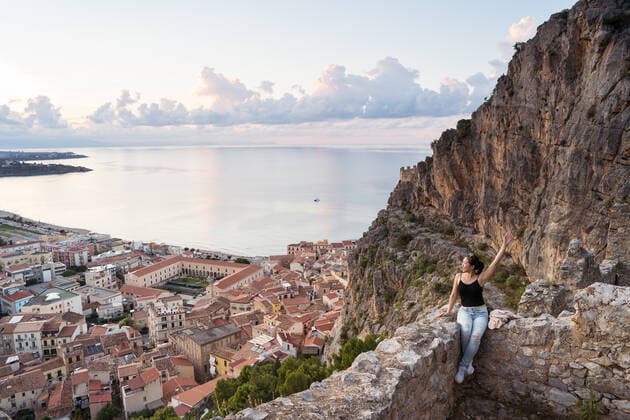 IMAGO Creative, Jahreszeit Sommer, Sommer in der Stadt Woman in casual clothes raising arm and admiring Cefalu City while sitting on old stone wall near mountain slope against sea and cloudy sunset sky in evening in Sicily, Italy JuanAlbertoRuiz_ID10183_308569_012 Copyright: xJuanxAlbertoxRuizx Model Released