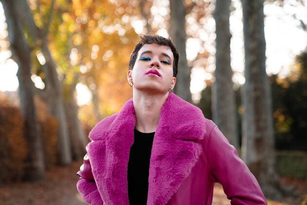 Self assured young gender fluid male with short hair and makeup in bright fuchsia leather and fur coat standing near autumn trees and looking at camera on sunny day, Model released DanielPerezLozao_ID10613_321516_002 Copyright: xDanielxPerezxLozaox MR