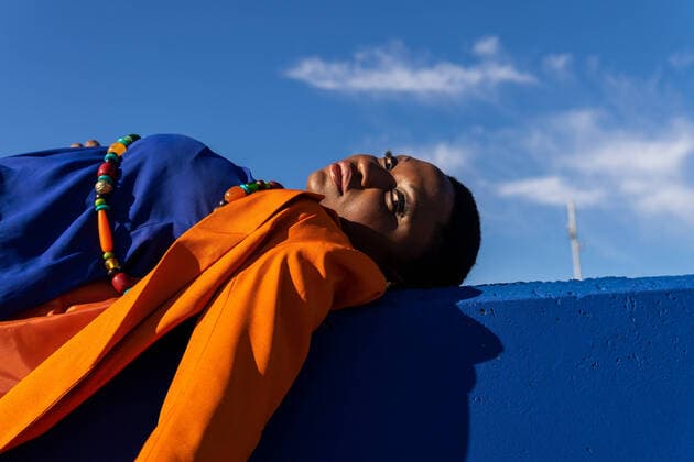 Creative Highlights Symbolbilder Low angle side view full body of African American female with short hair in orange suit and high heels lying on blue wall and enjoying sunny weather with blue sky looking at camera, Model released DavidMunoz_ID10670_323138_026 Copyright: xDavidxMunozx MR