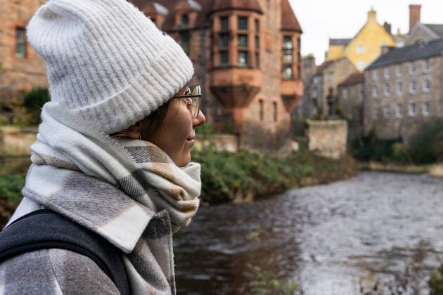 Happy young female in stylish outerwear and glasses smiling and looking away while standing on shore of narrow river on autumn day in Edinburgh, Scotland DavidMunoz_ID10673_323292_024 Copyright: xDavidxMunozx Model Released