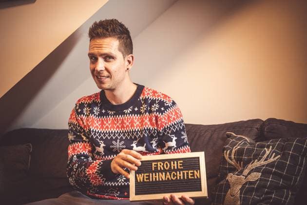Bavaria, Germany - 23 October 2022: Young man in Christmas sweater sits in living room at Christmas with a sign in hand with inscription: Merry Christmas *** Junger Mann in Weihnachtspullover sitzt im Wohnzimmer an Weihnachten mit einem Schild in der Hand mit Aufschrift: Frohe Weihnachten