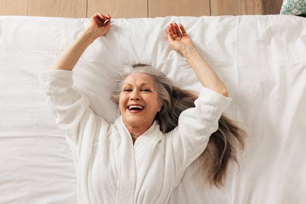 Creative Highlights Symbolbilder Happy senior woman in a bathrobe lying on a bed in a hotel room looking at camera Copyright: xArtemxVarnitsinxArtemxVarnitsinx