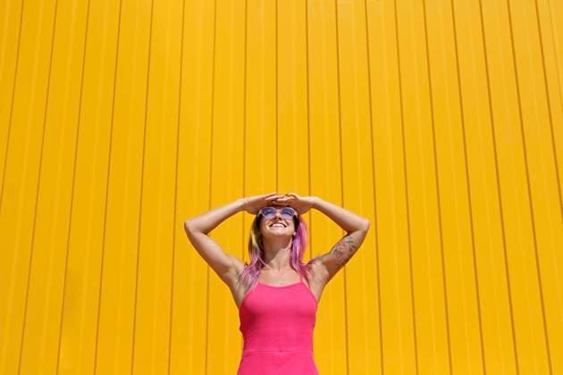 IMAGO Creative, Lifestyle, Barbiecore Happy woman wearing pink dress shielding eyes in front of yellow wall model released, Symbolfoto property released, SYEF00111