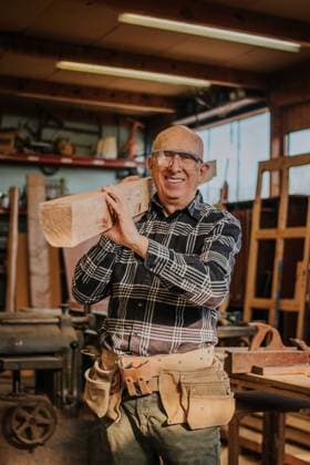 Smiling senior male carpenter in protective glasses carrying wood on shoulder in professional joinery during repair process, Model released DavidGari_ID11724_351231_043 Copyright: xDavidxGarix MR